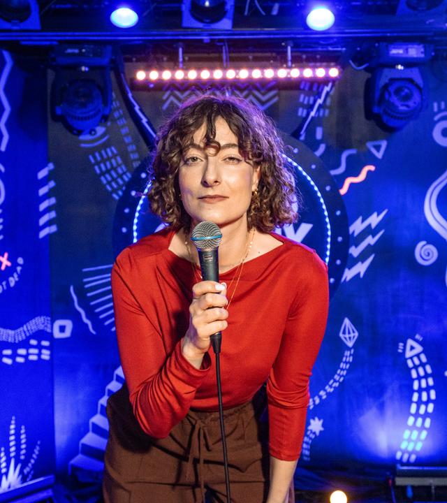 Lisa Frischemeier steht in der Mitte des Bildes und hat ein Mikrofon in der Hand. Der Hintergrund ist blau beleuchtet.