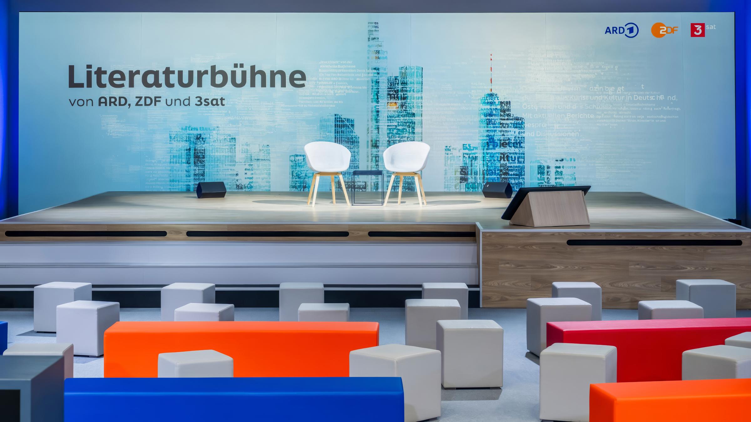 Literaturbühne von  ARD / ZDF / 3sat: Zwei weiße Stühle mit einem kleinen Tisch dazwischen stehen auf einer Bühne. Im Hintergrund linke der Schriftzug Literaturbühne von  ARD / ZDF / 3sat. Oben rechts Logos von ARD, ZDF, 3sat auf grauem Hintergrund. Vor der Bühne stehen Sitzhocker und Bänke in den Farben weiß, blau, orange und rot. 