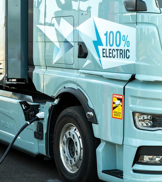 Nahaufnahme eines vollelektrischen Lastwagens beim Laden. Rechts füllt das hellblaue Führerhaus den größten Teil des Bildes aus, links steht eine hohe Ladesäule. Das angeschlossene Kabel verläuft über den Boden zum Lkw; die Aufschrift „100 % Electric“ weist auf dessen batterieelektrischen Antrieb hin.
