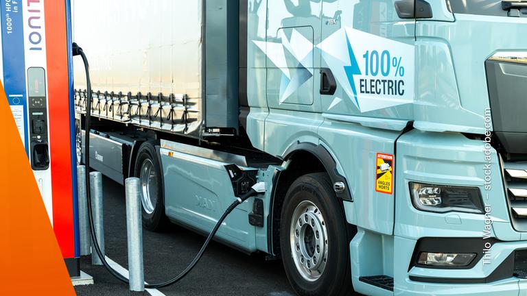 Nahaufnahme eines vollelektrischen Lastwagens beim Laden. Rechts füllt das hellblaue Führerhaus den größten Teil des Bildes aus, links steht eine hohe Ladesäule. Das angeschlossene Kabel verläuft über den Boden zum Lkw; die Aufschrift „100 % Electric“ weist auf dessen batterieelektrischen Antrieb hin.