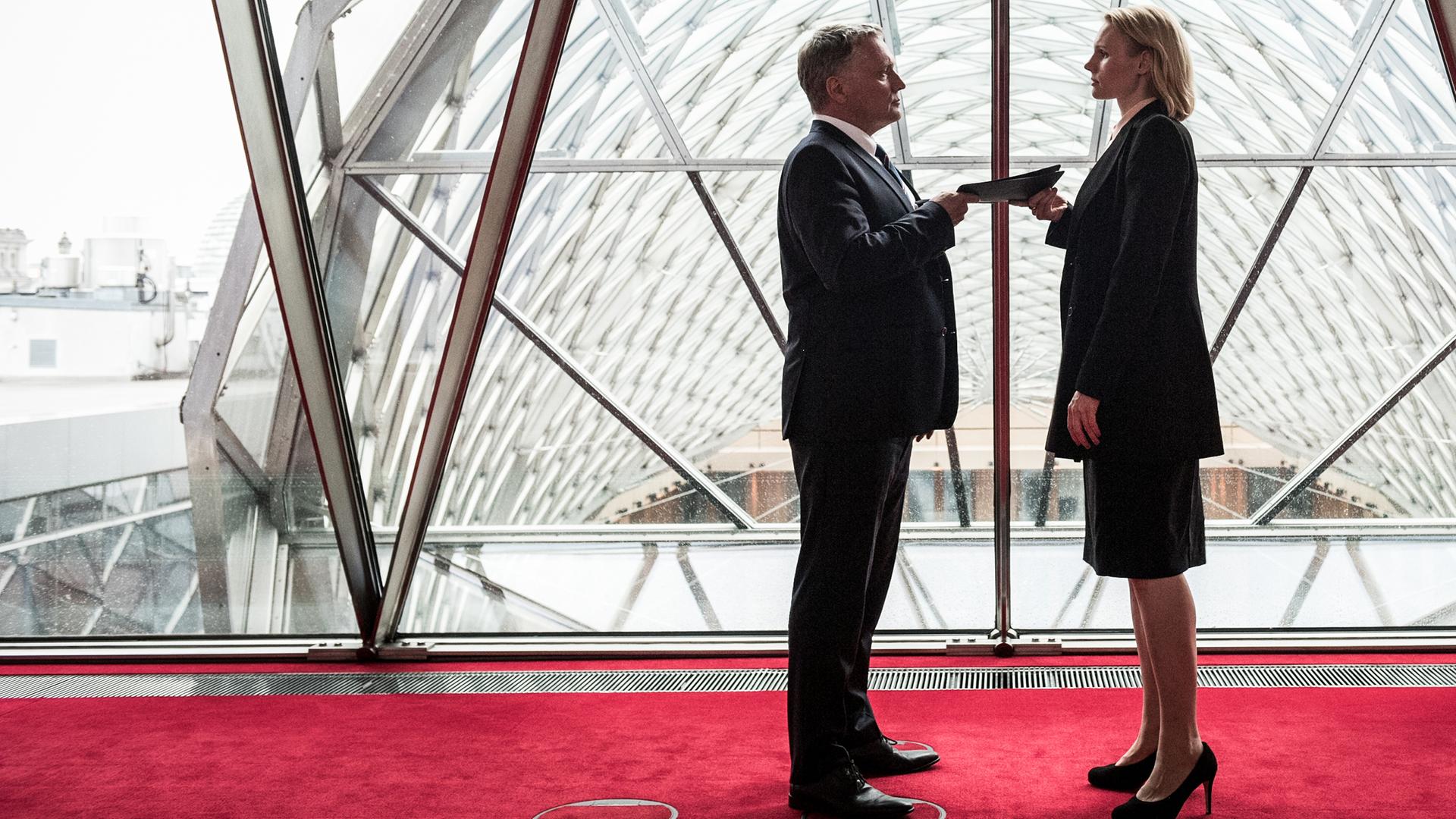 Eva Blumenthal (Rosalie Thomass) steht Wolfgang Zielert (Bernhard Schir) auf einem roten Teppich vor einer Glasfront gegenüber und übergibt ihm ein Schriftstück.