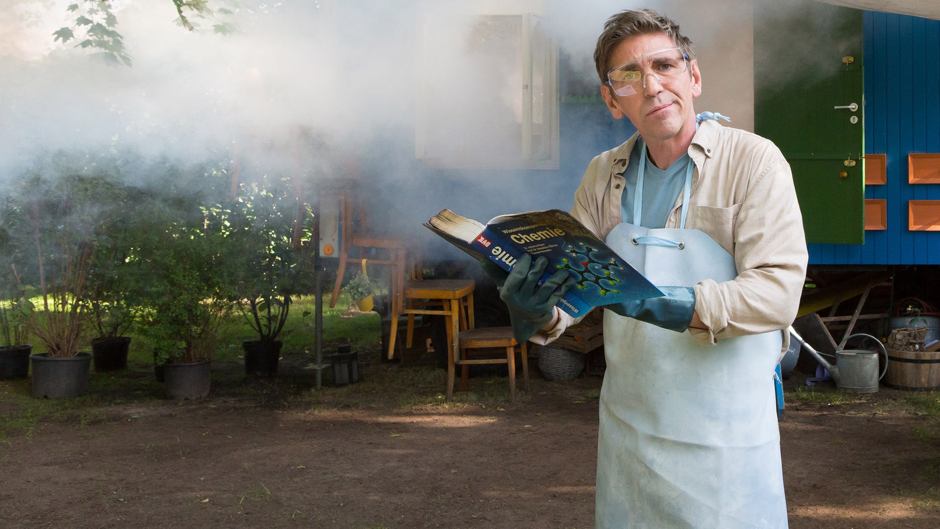 Fritz steht mit einer Schutzmaske und einem Chemiebuch vor dem Bauwagen, der in Rauch eingehüllt ist.