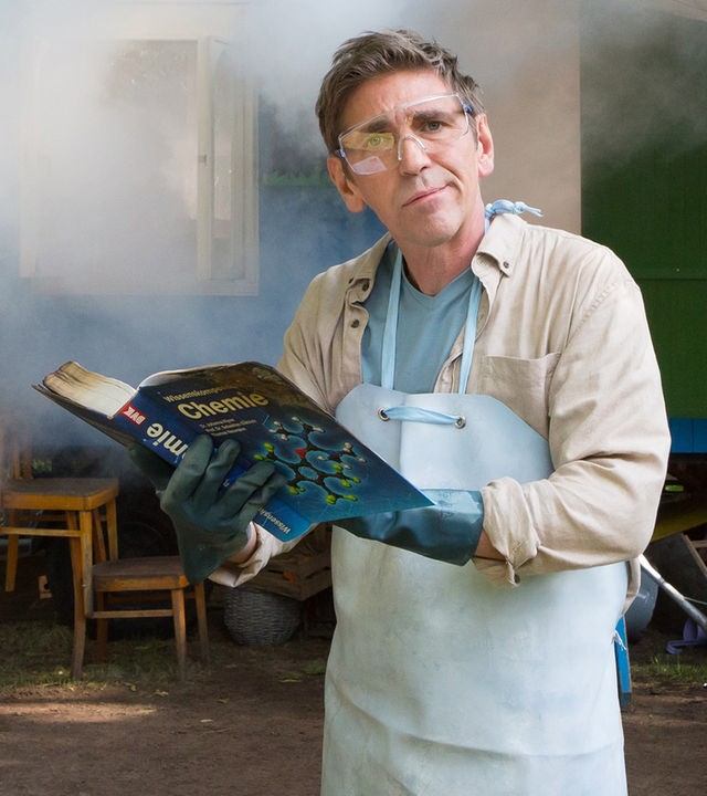 Fritz steht mit einer Schutzmaske und einem Chemiebuch vor dem Bauwagen, der in Rauch eingehüllt ist.