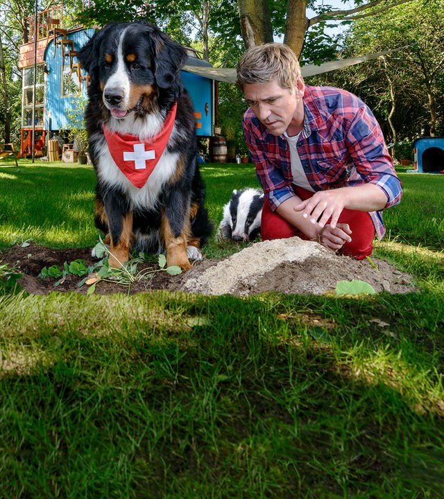 Fritz Fuchs hockt mit Keks auf dem Rasen vor einem Dachsbau. Der Dachs sitzt zwischen den Beiden.