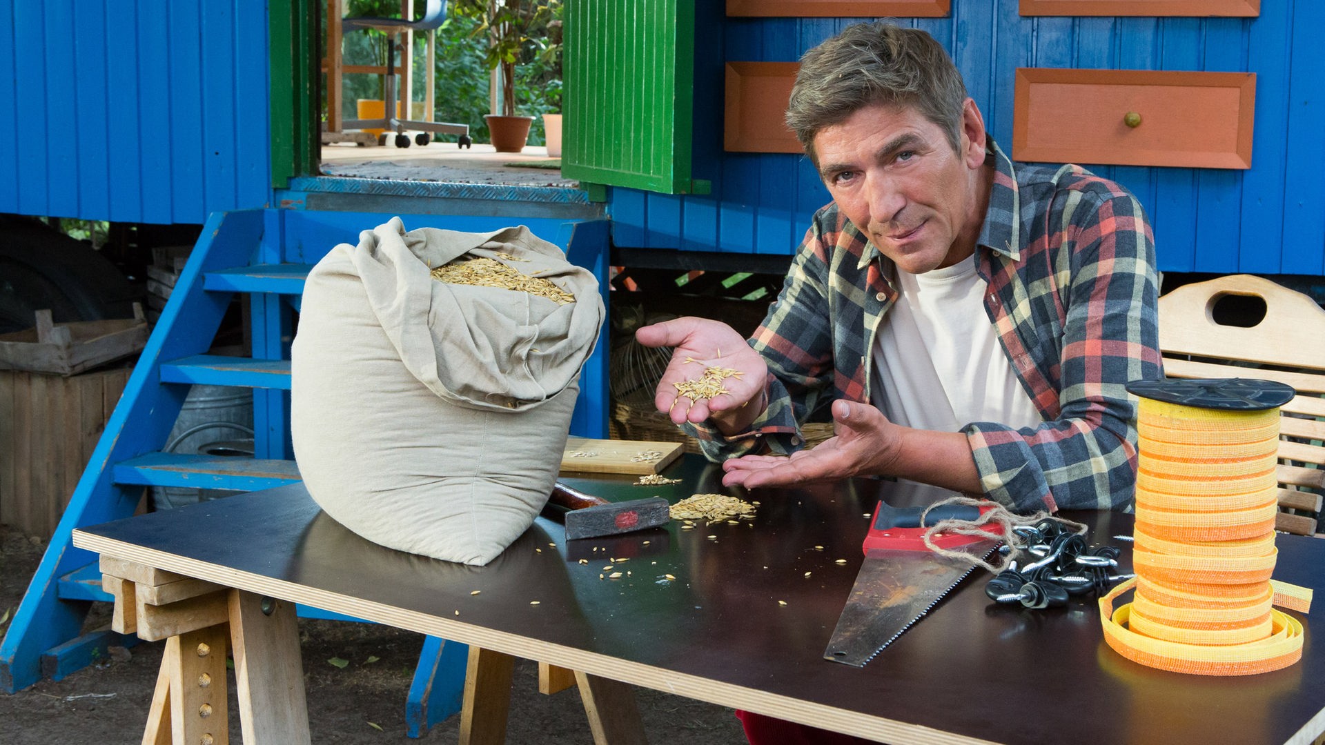 Fritz sitzt an einem Tisch vor dem Bauwagen und hält Haferkörner in seinen Händen.
