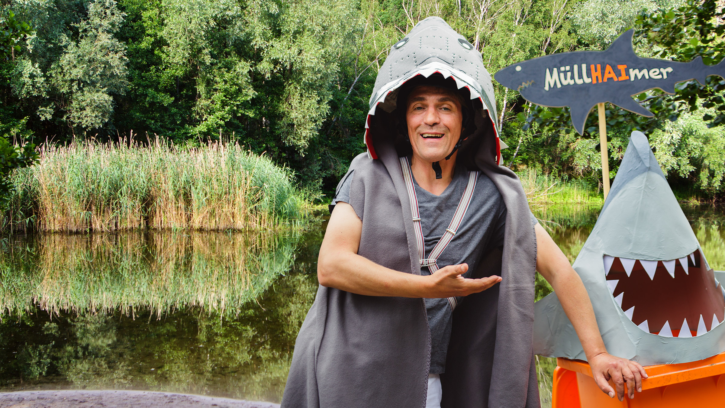 Fritz steht mit einem Haikostüm verkleidet am Seeufer, neben ihm eine Mülltonne als Hai