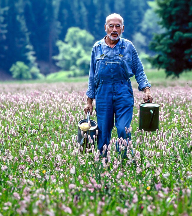 Peter Lustig geht über eine Wiese und hat in jeder Hand eine Gießkanne