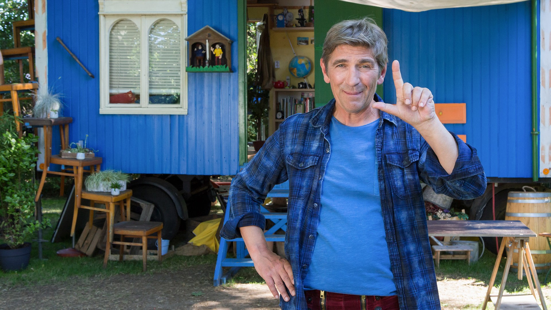 Fritz steht vor dem Bauwagen und zeigt mit den Fingern der linken Hand ein L