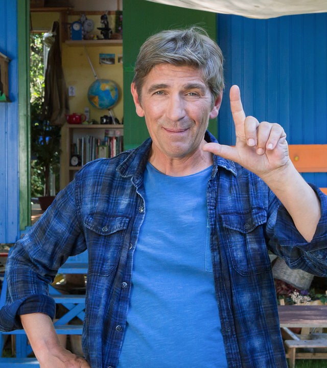 Fritz steht vor dem Bauwagen und zeigt mit den Fingern der linken Hand ein L