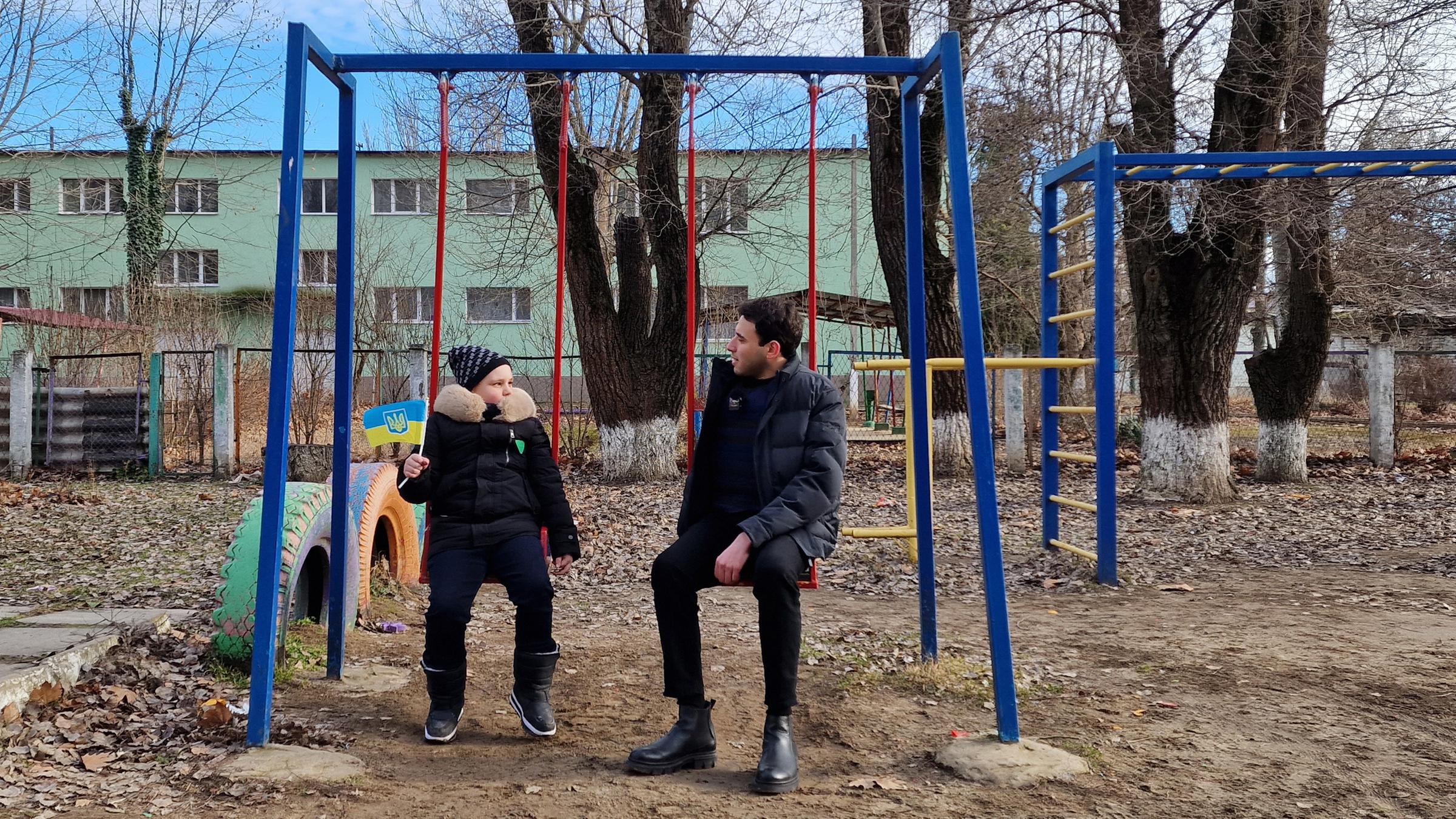  logo! extra: Kinder in der Ukraine - ein Jahr im Krieg: Moderator Sherif Rizkallah und ein Kind  sitzen auf Schaukeln nebeneinander und unterhalten sich.