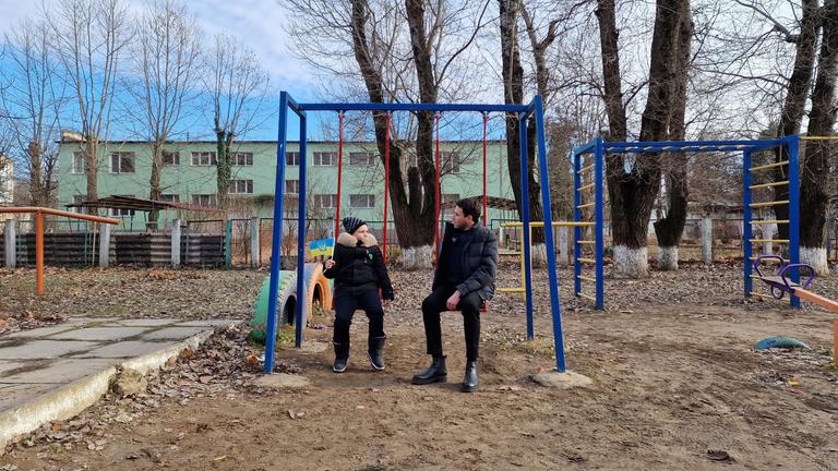  logo! extra: Kinder in der Ukraine - ein Jahr im Krieg: Moderator Sherif Rizkallah und ein Kind  sitzen auf Schaukeln nebeneinander und unterhalten sich.