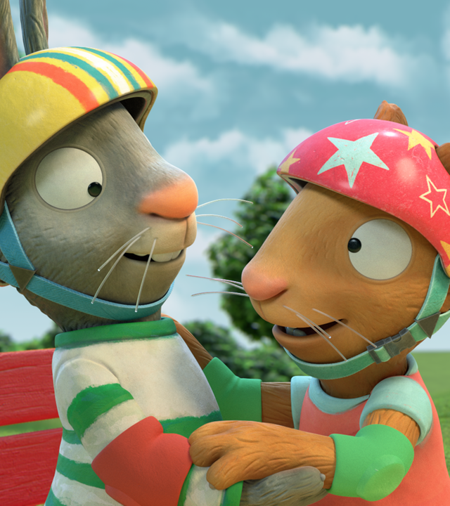 Pip und Posy umarmen sich und haben einen Fahrradhelm auf. Pips Helm ist gelb und ist bunt gestreift, Posys Helm ist rot und hat gelbe und weisse Sterne.
