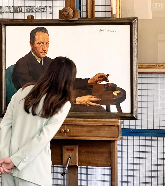 Eine Frau mit langen, dunkelbraunen Haaren steht mit dem Rücken zur Kamera und betrachtet ein Gemälde. Sie trägt einen hellen Hosenanzug und hat die Hände locker hinter ihrem Rücken verschränkt. Das Gemälde zeigt einen Mann in dunkler Kleidung, der an einem kleinen runden Tisch sitzt und eine Zigarre in der Hand hält. Neben ihm auf dem Tisch liegen weitere Gegenstände, darunter ein Streichholzbriefchen. Der Rahmen des Bildes ist aus dunklem Holz. Im Hintergrund sind weitere leere Bilderrahmen sowie ein Metallgitter zu sehen. 