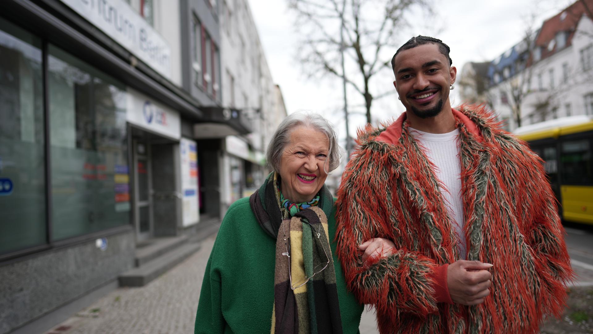 Louis (rechts) und seine Oma (links) schauen lächelnd in die Kamera. Sie hat sich bei ihm am Arm eingehakt. Im Hintergrund sieht man einen Baum und eine Straße.