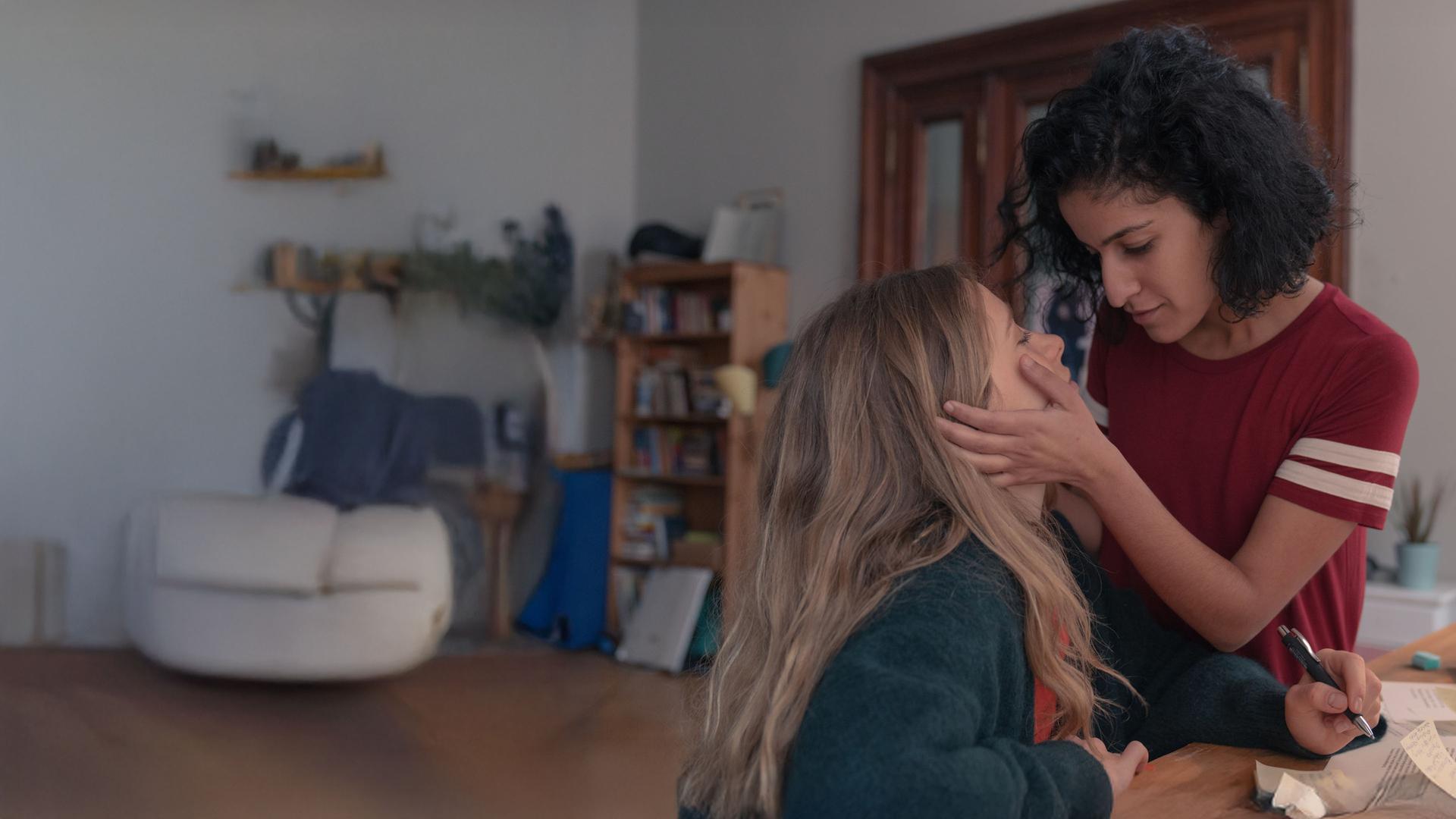 "Loving Her - Loving Franzi": Franzi (Lena Klenke) sitzt an einem Schreibtisch. Vor ihr liegen Papiere. In ihrer linken Hand hält sie einen Kugelschreiber. Hanna (Banafshe Hourmazdi) steht dicht neben ihr. Sie hat ihre Hände auf Franzis Wangen liegen. Beide sehen sich verliebt an.