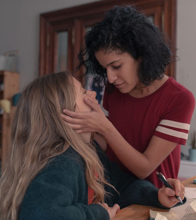 "Loving Her - Loving Franzi": Franzi (Lena Klenke) sitzt an einem Schreibtisch. Vor ihr liegen Papiere. In ihrer linken Hand hält sie einen Kugelschreiber. Hanna (Banafshe Hourmazdi) steht dicht neben ihr. Sie hat ihre Hände auf Franzis Wangen liegen. Beide sehen sich verliebt an.