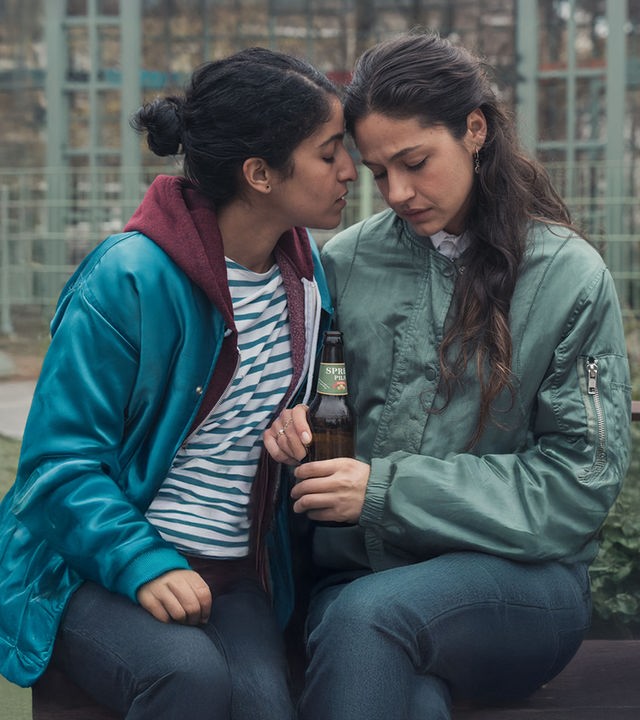 "Loving Her - Loving Sarah": Hanna (Banafshe Hourmazdi) und Sarah (Soma Pysall) sitzen eng nebeneinander. Hanna hat ihren Kopf zu Sarah gedreht. Sarah blickt nach unten. Sie hält in ihrer rechten Hand eine Flasche Bier.