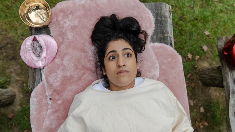 "Loving Her": Großaufnahme von Hanna (Banafshe Hourmazdi). Sie liegt auf einem mit rosa Plüschdecken gepolsterten Holztisch. Neben ihrem Kopf steht eine Klangschale. Daneben liegt ein rosa Beutel. Sie hat die Augen weit geöffnet und blickt fragend.