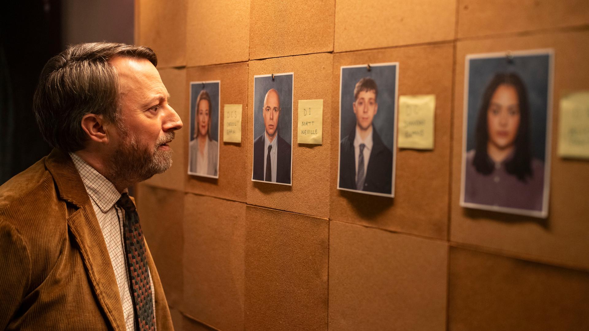 "Ludwig - Ein Hilferuf aus Cambridge": John Taylor (David Mitchell) steht vor einer großen Pinnwand, an der einige ausgedruckte Porträtfotos und kleine Zettel mit Infos hängen, und betrachtet eines der Bilder genau.