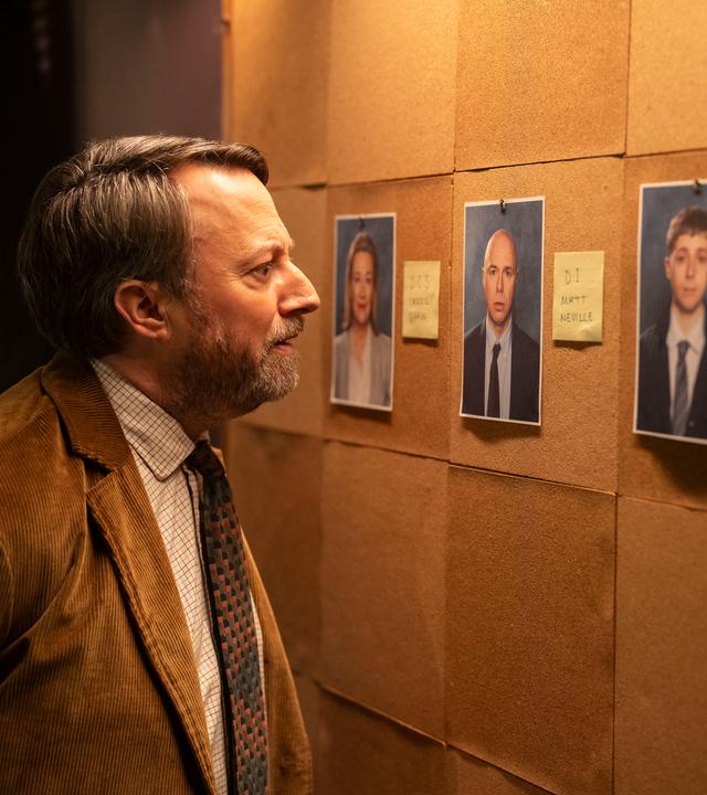 "Ludwig - Ein Hilferuf aus Cambridge": John Taylor (David Mitchell) steht vor einer großen Pinnwand, an der einige ausgedruckte Porträtfotos und kleine Zettel mit Infos hängen, und betrachtet eines der Bilder genau.