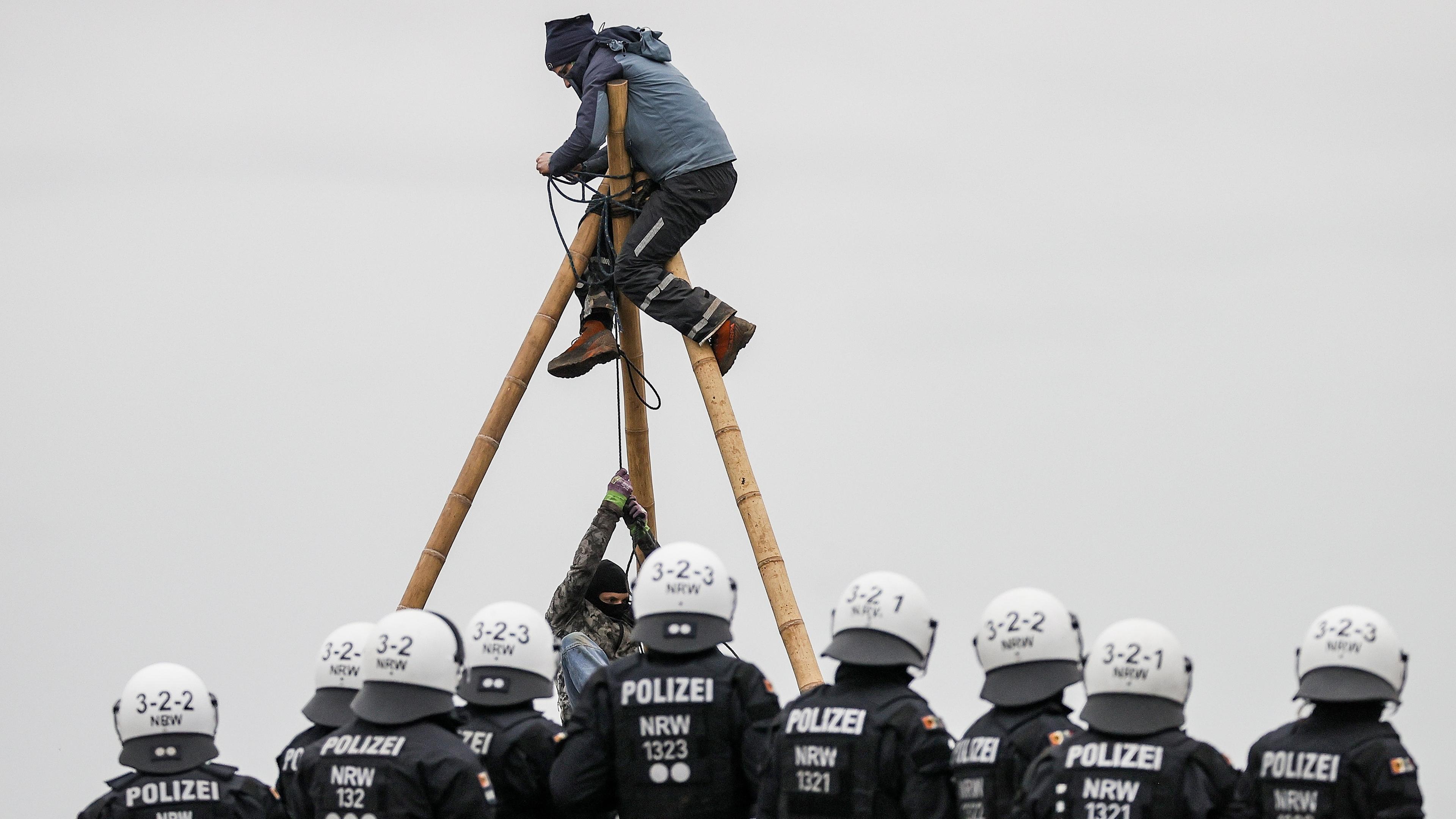 Aktivisten hängen in einem Stativ nahe Lützeraht, um die Räumung des Orts zu verhindern. Polizisten beobachten die Situation.