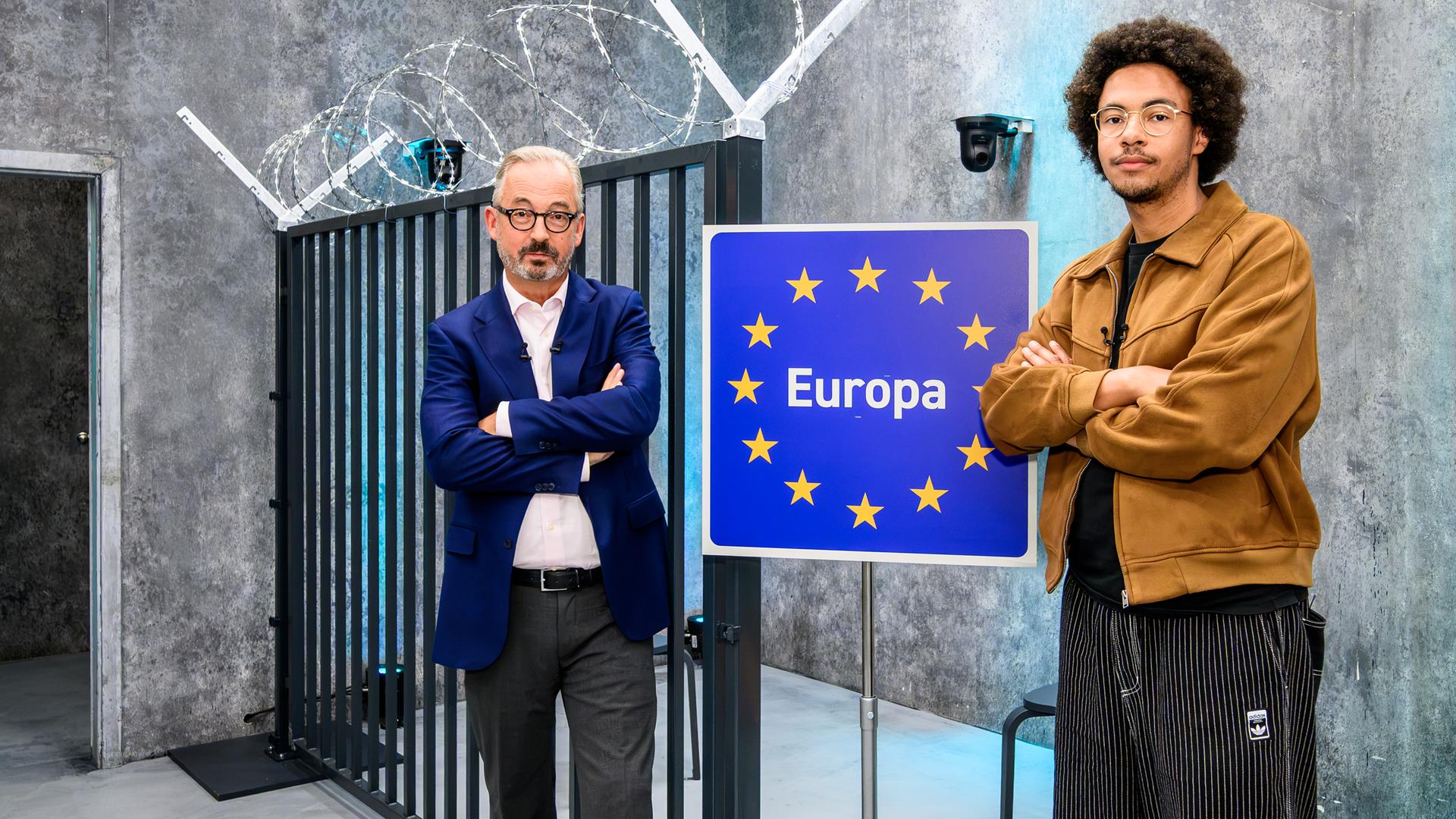 Jan Fleischhauer und Luis Bobga stehen mit verschränkten Armen vor einem schwarzen Metalltor im Studio. Zwischen beiden steht ein blaues Schild mit gelben Sternen und der Aufschrift Europa. Über dem Tor ist spiralförmig Stacheldraht angebracht.