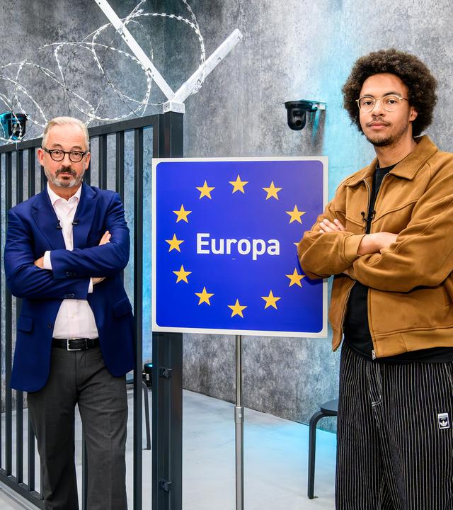 Jan Fleischhauer und Luis Bobga stehen mit verschränkten Armen vor einem schwarzen Metalltor im Studio. Zwischen beiden steht ein blaues Schild mit gelben Sternen und der Aufschrift Europa. Über dem Tor ist spiralförmig Stacheldraht angebracht.