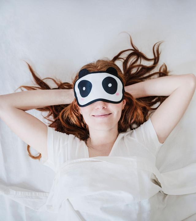Eine Frau liegt auf dem Rücken auf einem weißen Bettlaken. Sie hat eine Pandabären-Schlafmaske auf.