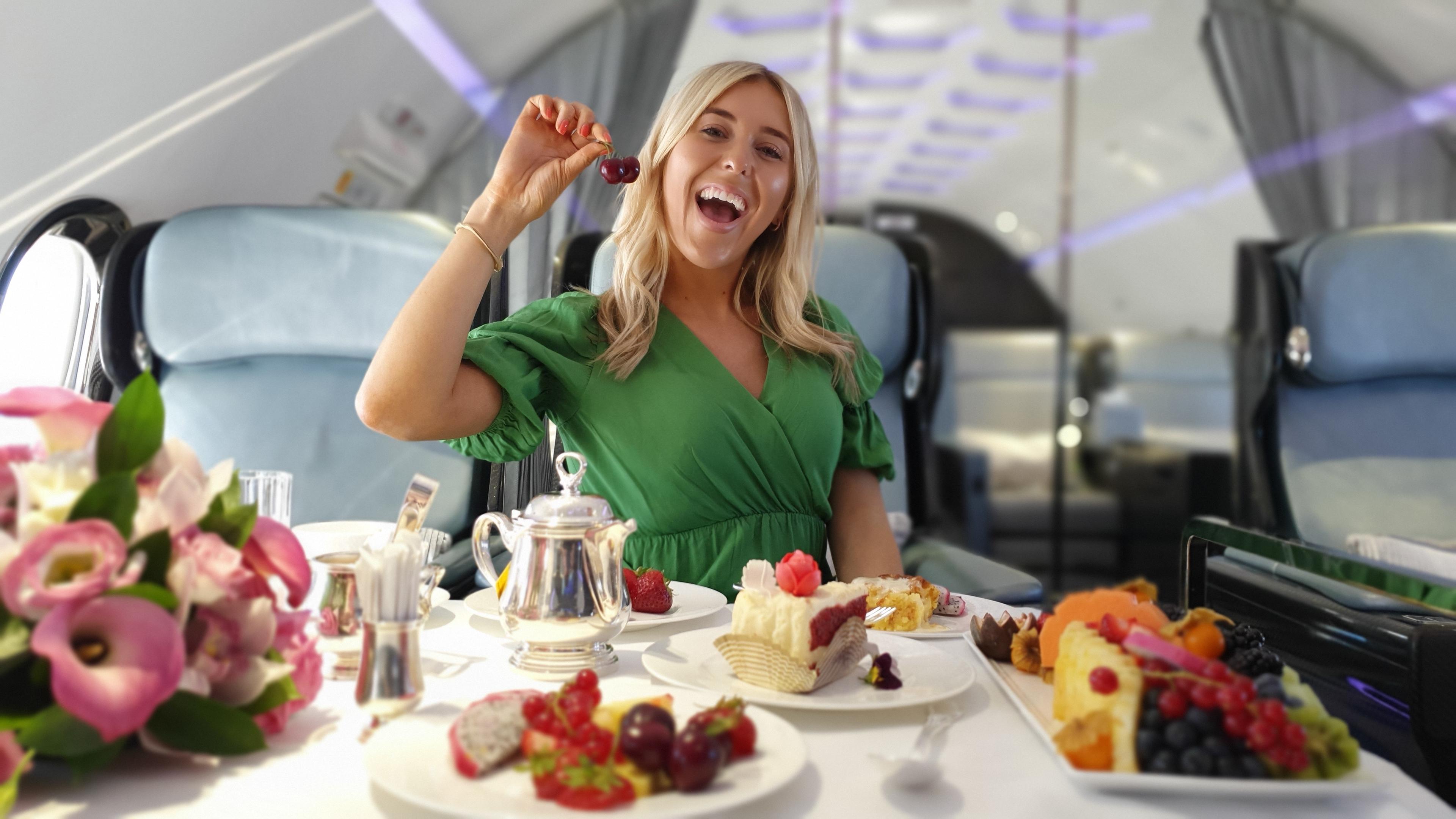 Luxusbloggerin Em Sheldon sitzt in einem Privat-Jet vor einem gedeckten Tisch und isst Kirschen.