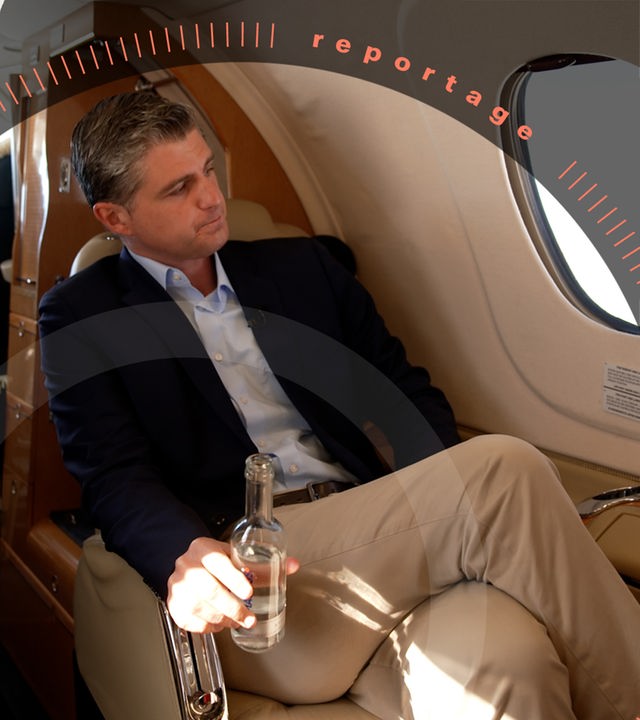 Julien Backhaus sitzt mit überschlagenen Beinen in einem Privatjet mit einer Wasserflasche in der Hand. Er schaut aus dem Fenster. Im Hintergrund sind die Schultern von Pilot und Co-Pilot im Cockpit zu sehen.