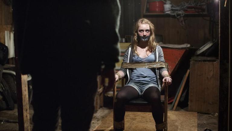 Lykke (Sara Månberg) ist mit Klebeband an einen Stuhl gefesselt und schaut einen Mann an, der ihr gegenüber sitzt. Ihr Mund ist ebenfalls mit Klebeband beklebt.