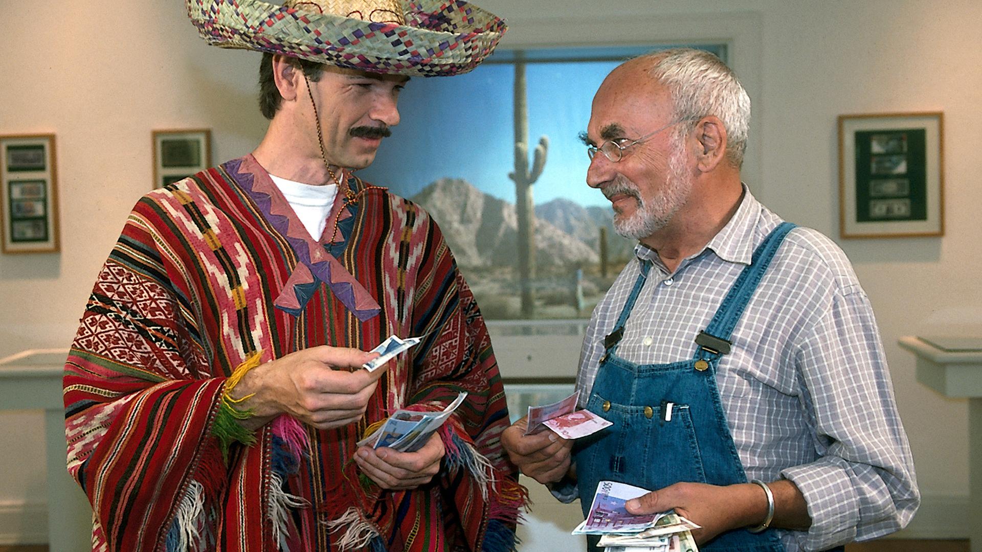 Peter steht in einem Ausstellungsraum neben einem Mann mit Sombrero, Poncho und Schnurrbart. Beide haben einige Geldscheine in der Hand und lächeln sich an.