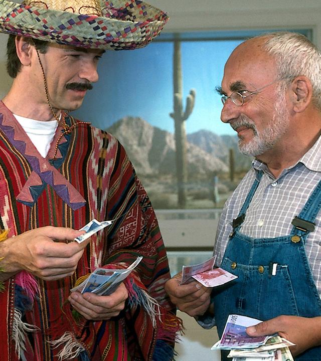 Peter steht in einem Ausstellungsraum neben einem Mann mit Sombrero, Poncho und Schnurrbart. Beide haben einige Geldscheine in der Hand und lächeln sich an.