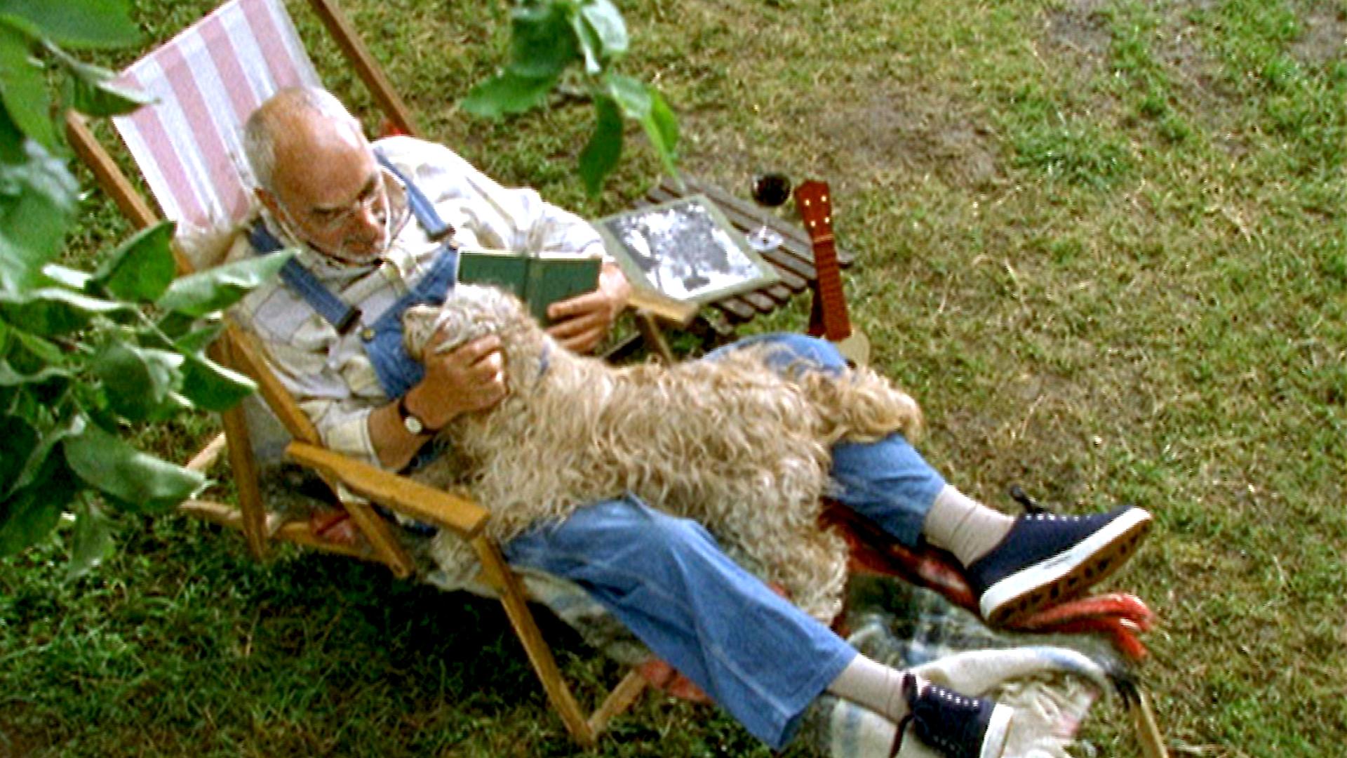 Bild aus der Vogelperspektive: Peter sitzt im Garten auf einem Stuhl. Er hat ein Buch in der einen Hand und krault mit der anderen einen weißen Hund, der auf ihm liegt.