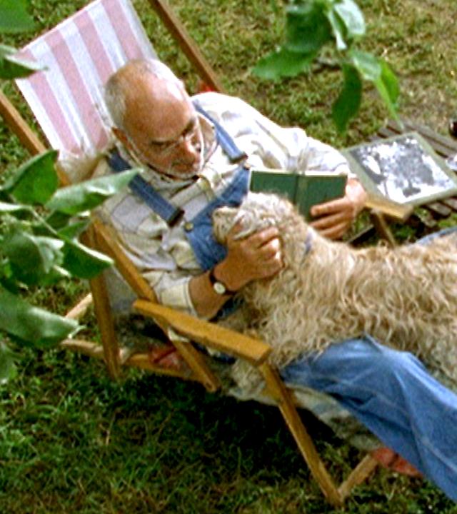 Bild aus der Vogelperspektive: Peter sitzt im Garten auf einem Stuhl. Er hat ein Buch in der einen Hand und krault mit der anderen einen weißen Hund, der auf ihm liegt.