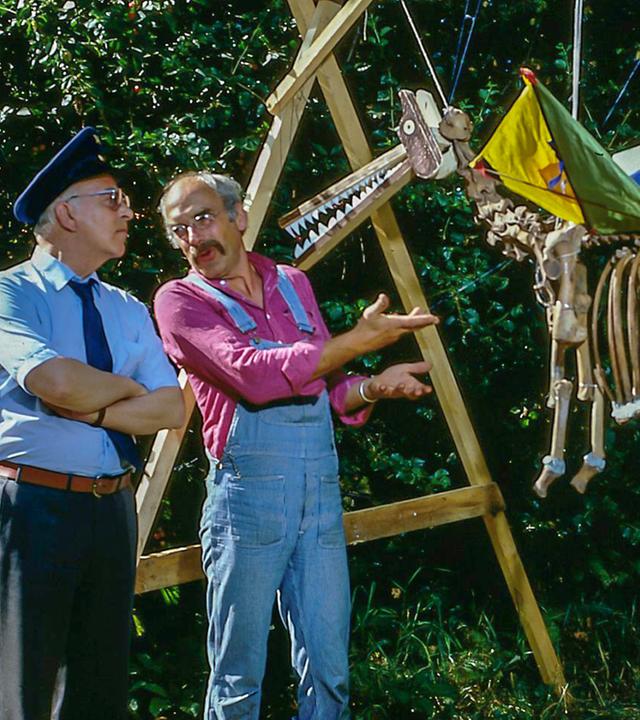 Peterlustig erklärt einem Polizisten ein aufgehängtes Tierskelett. Das Tierskelett hat durch zwei Drachen Flügel bekommen und erinnert so selbst an einen Drachen. 