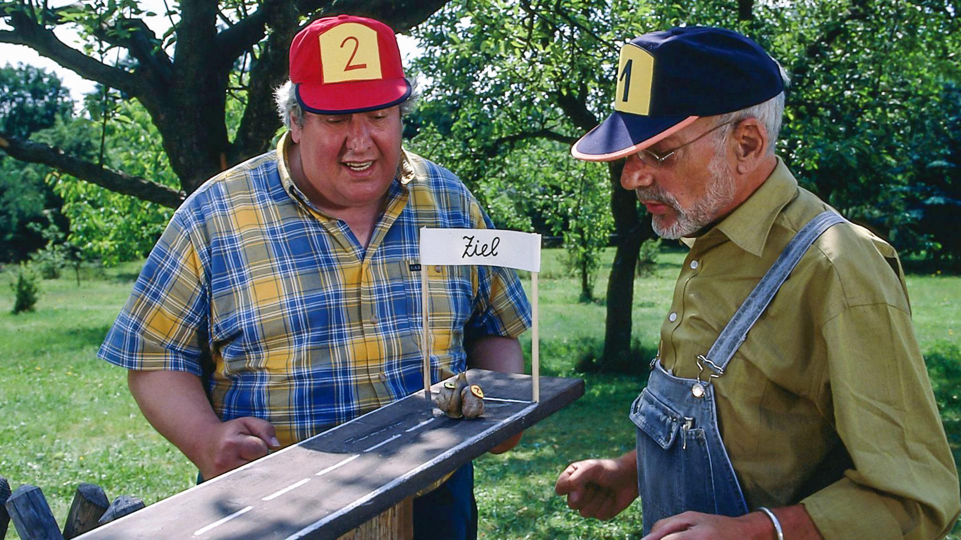 Peter Lustig und Nachbar Paschulke haben im Freien eine Miniatur-Rennbahn aufgebaut, die in der Gestaltung einer ansphaltierten Straße ähnelt und die sie gespannt beobachten. Zwei Schnecken, deren Häuser mit den Nummern 1 und 2 markiert sind, kriechen kurz vor der Zielgeraden. Peter trägt eine Mütze mit der Nummer 1, Paschulke eine mit der Nummer 2.