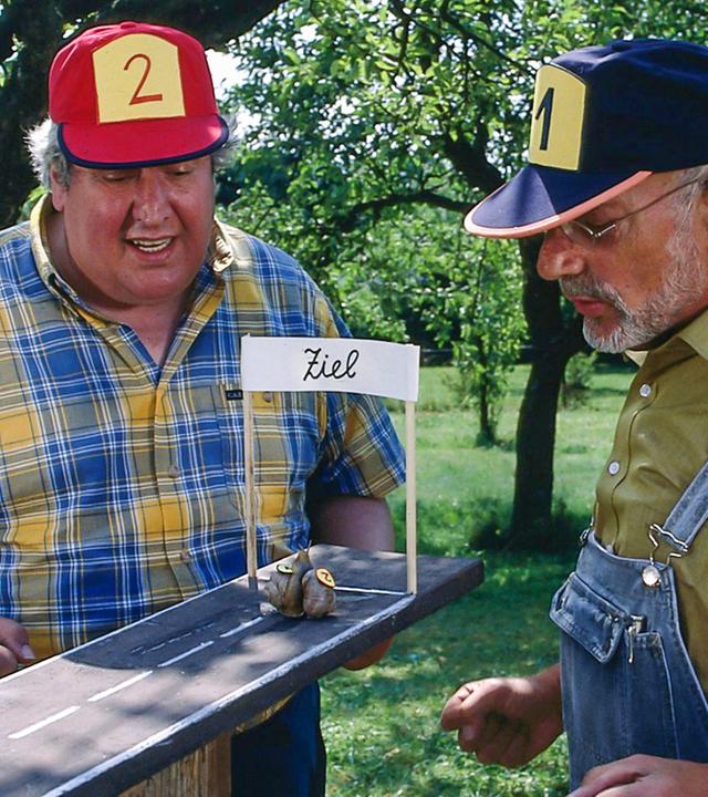 Peter Lustig und Nachbar Paschulke haben im Freien eine Miniatur-Rennbahn aufgebaut, die in der Gestaltung einer ansphaltierten Straße ähnelt und die sie gespannt beobachten. Zwei Schnecken, deren Häuser mit den Nummern 1 und 2 markiert sind, kriechen kurz vor der Zielgeraden. Peter trägt eine Mütze mit der Nummer 1, Paschulke eine mit der Nummer 2.