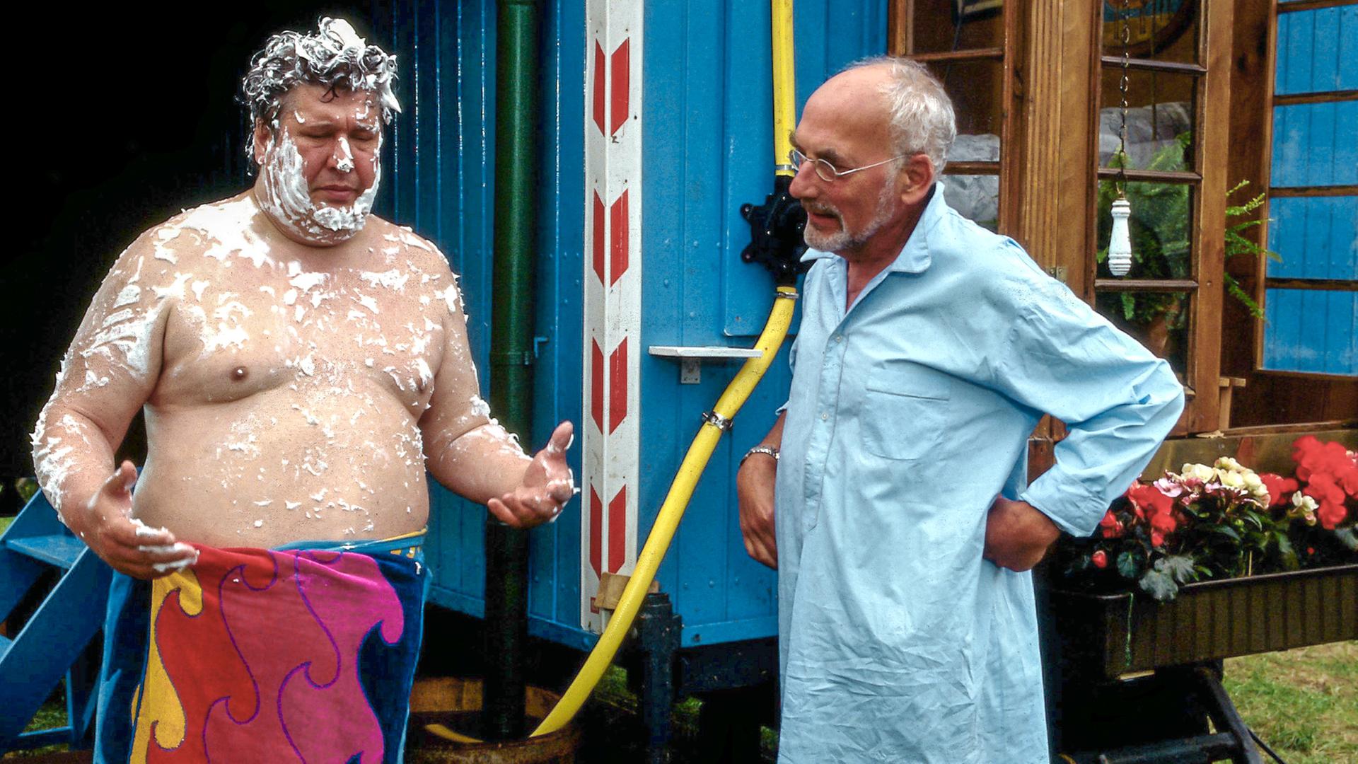 Peter Lustig steht im Nachthemd rechts vor seinem Wohnwagen. Links steht Nachbar Paschulke. Er trägt nur ein Handtuch um die Hüfte und ist an Oberkörper und Kopf mit Seife bedeckt.