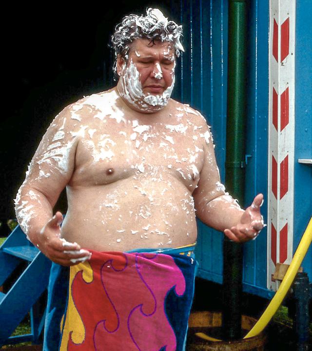 Peter Lustig steht im Nachthemd rechts vor seinem Wohnwagen. Links steht Nachbar Paschulke. Er trägt nur ein Handtuch um die Hüfte und ist an Oberkörper und Kopf mit Seife bedeckt.