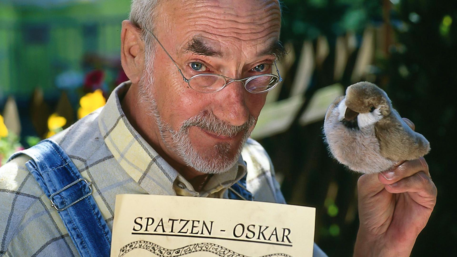 Peter Lustig in der Nahaufnahme: Er schaut in die Kamera, in seiner linken Hand hält er einen Plüsch-Vogel. In der anderen Hand hält er ein Papier mit dem Text "Spatzen-Oskar".