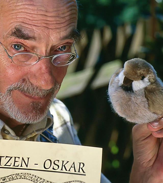 Peter Lustig in der Nahaufnahme: Er schaut in die Kamera, in seiner linken Hand hält er einen Plüsch-Vogel. In der anderen Hand hält er ein Papier mit dem Text "Spatzen-Oskar".
