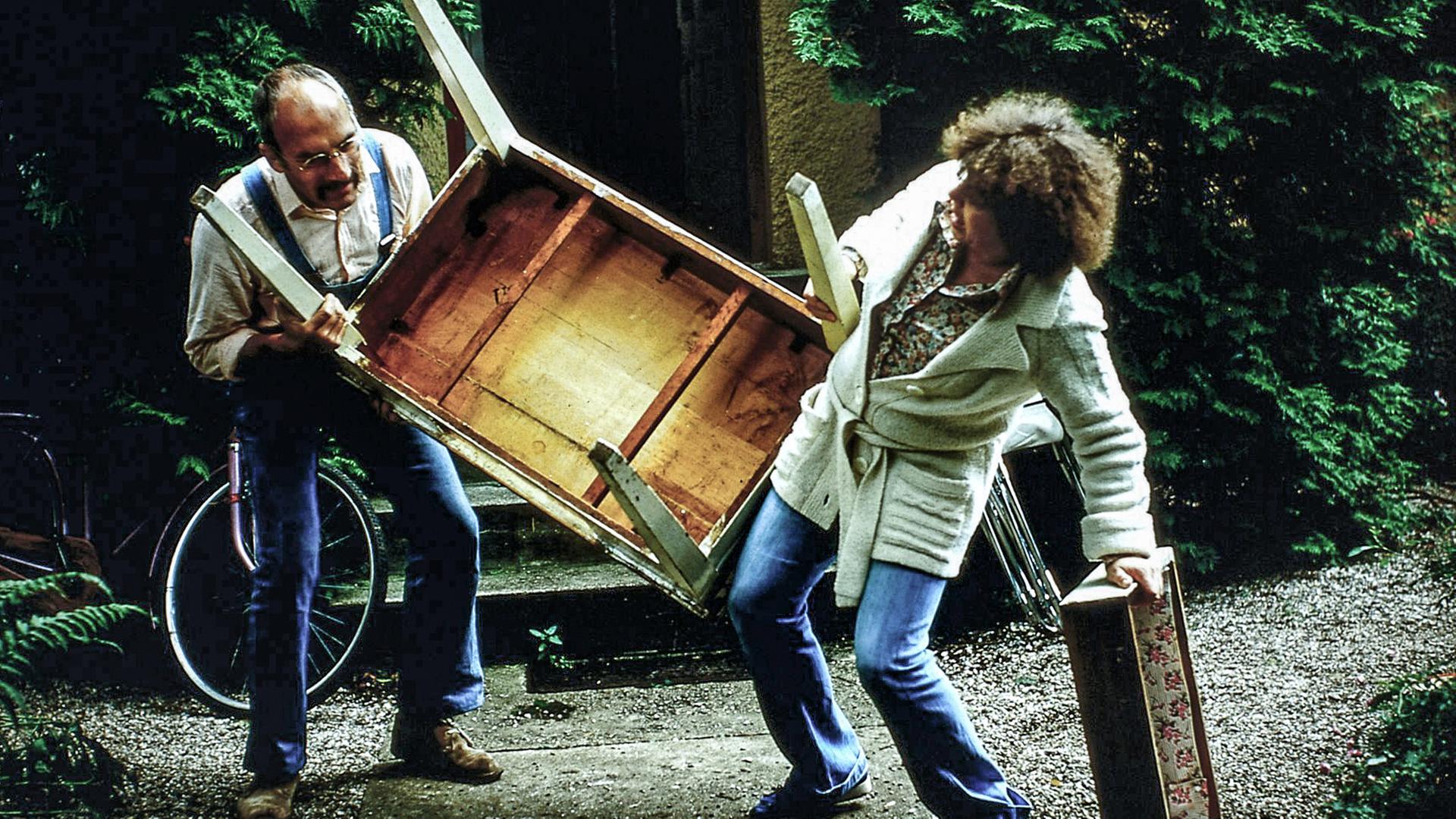 Peter Lustig trägt zusammen mit einer Frau einen schweren Tisch. Beide machen dabei eine krumme Figur.