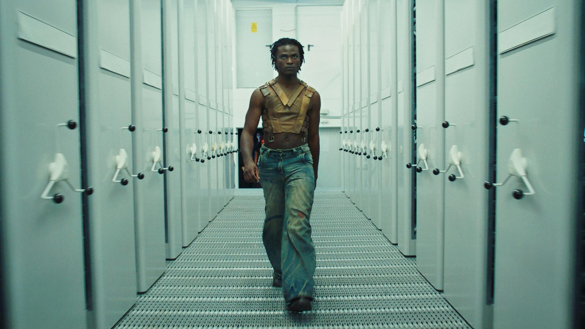  Das Bild zeigt eine Person, die selbstbewusst einen Korridor entlanggeht. Der Korridor ist von großen, grauen Schränken gesäumt, die auf beiden Seiten stehen. Die Person trägt ein bauchfreies, ärmelloses Oberteil aus braunem Leder mit Riemen und eine weit geschnittene Jeans. Das Licht ist gedämpft und erzeugt eine kühle Atmosphäre, die durch die grauen Wände und den metallenen Boden verstärkt wird. Die Person schaut direkt nach vorne, während sie auf die Kamera zugeht. 