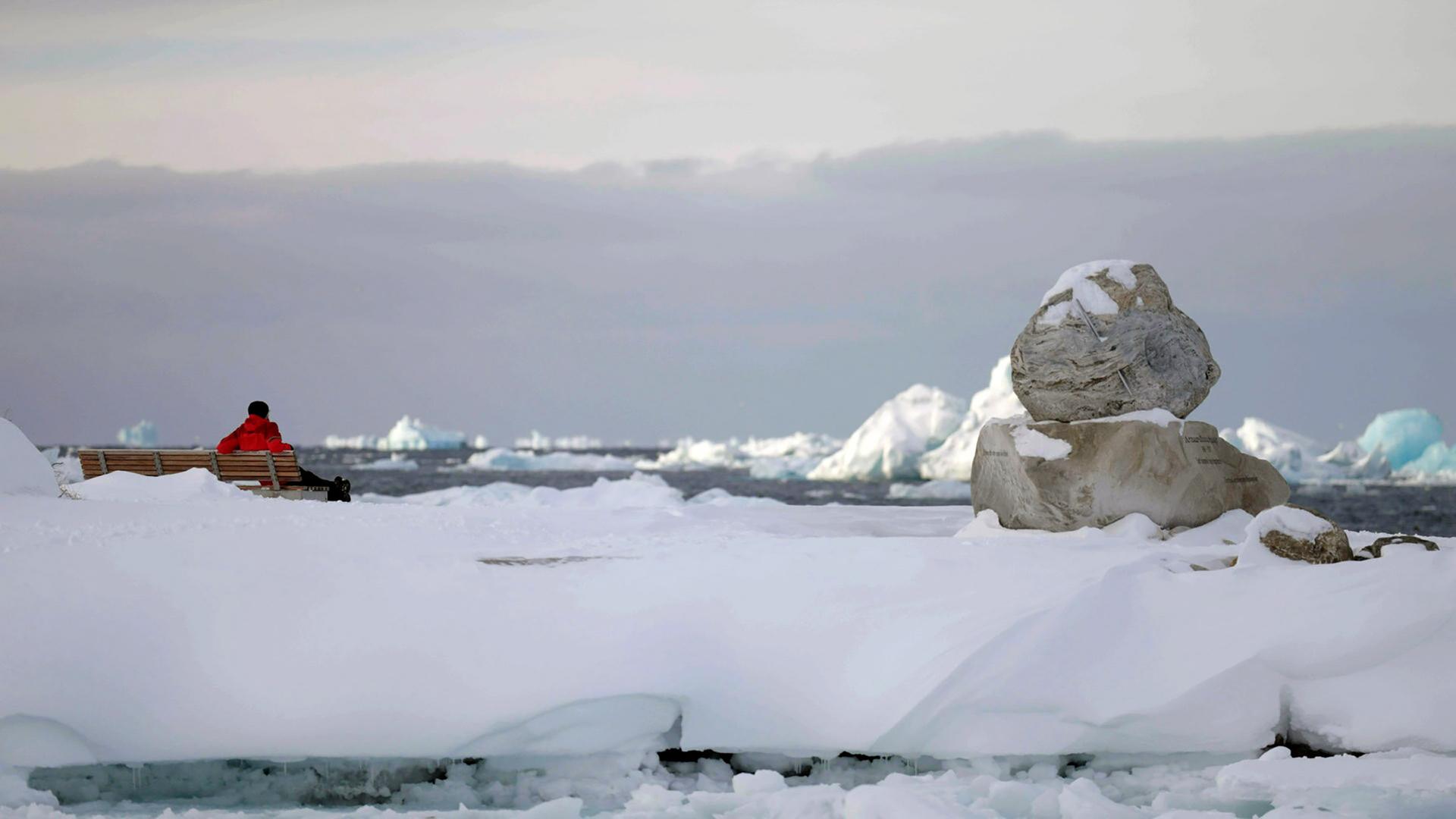 Eine Holzbank, auf der ein Mensch in roter Jacke sitzt und auf eine Wasserfläche mit vielen Eisbergen schaut.