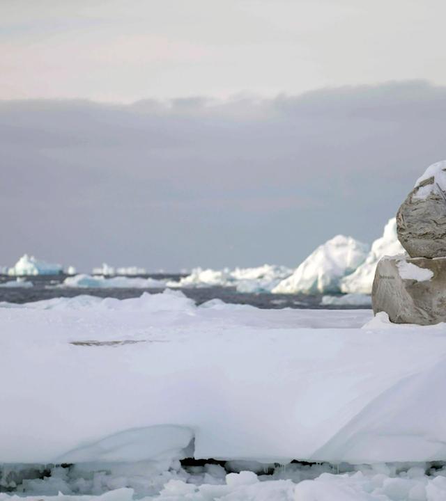 Eine Holzbank, auf der ein Mensch in roter Jacke sitzt und auf eine Wasserfläche mit vielen Eisbergen schaut.