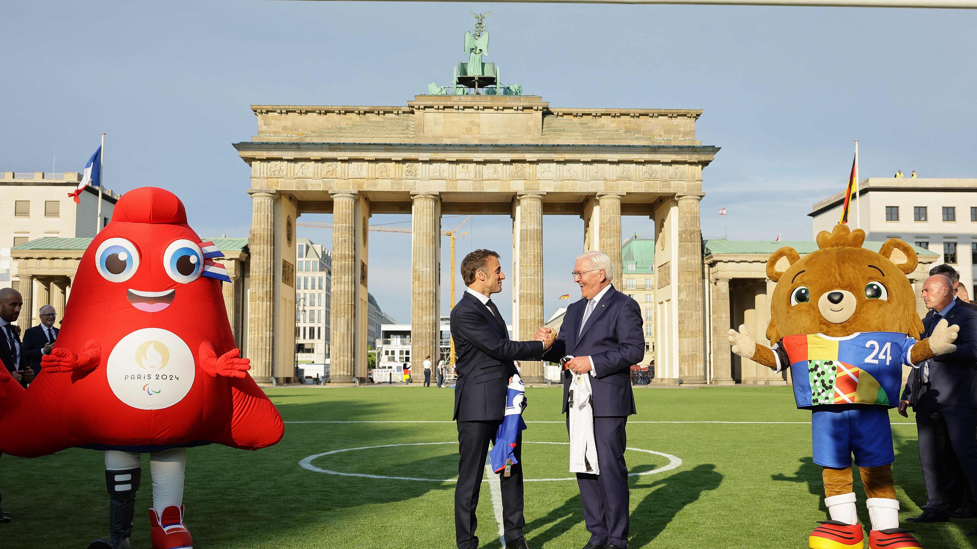 French President Emmanuel Macron and German President Frank-Walter Steinmeier shake hands as they exchance football shirts next to "Albaert" the official mascot of the UEFA Euro 2024 European Football Championship (R) and "Phryge" the Paris 2024 Olympic mascot, as they inaugurate the German-French Sports Summer 2024 in front of the Brandenburg Gate in Berlin, Germany on May 26, 2024. 