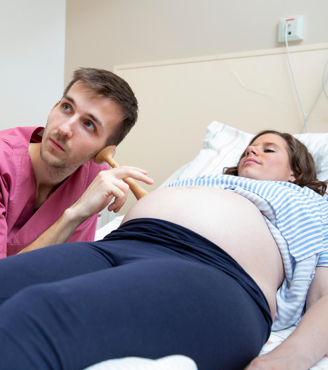 Casper Jaginiak, ein junger Mann in Krankenhauskleidung, hört mit einem Hörrohr (Stethoskop) den Bauch einer Frau ab, die auf einer Liege vor ihm liegt.