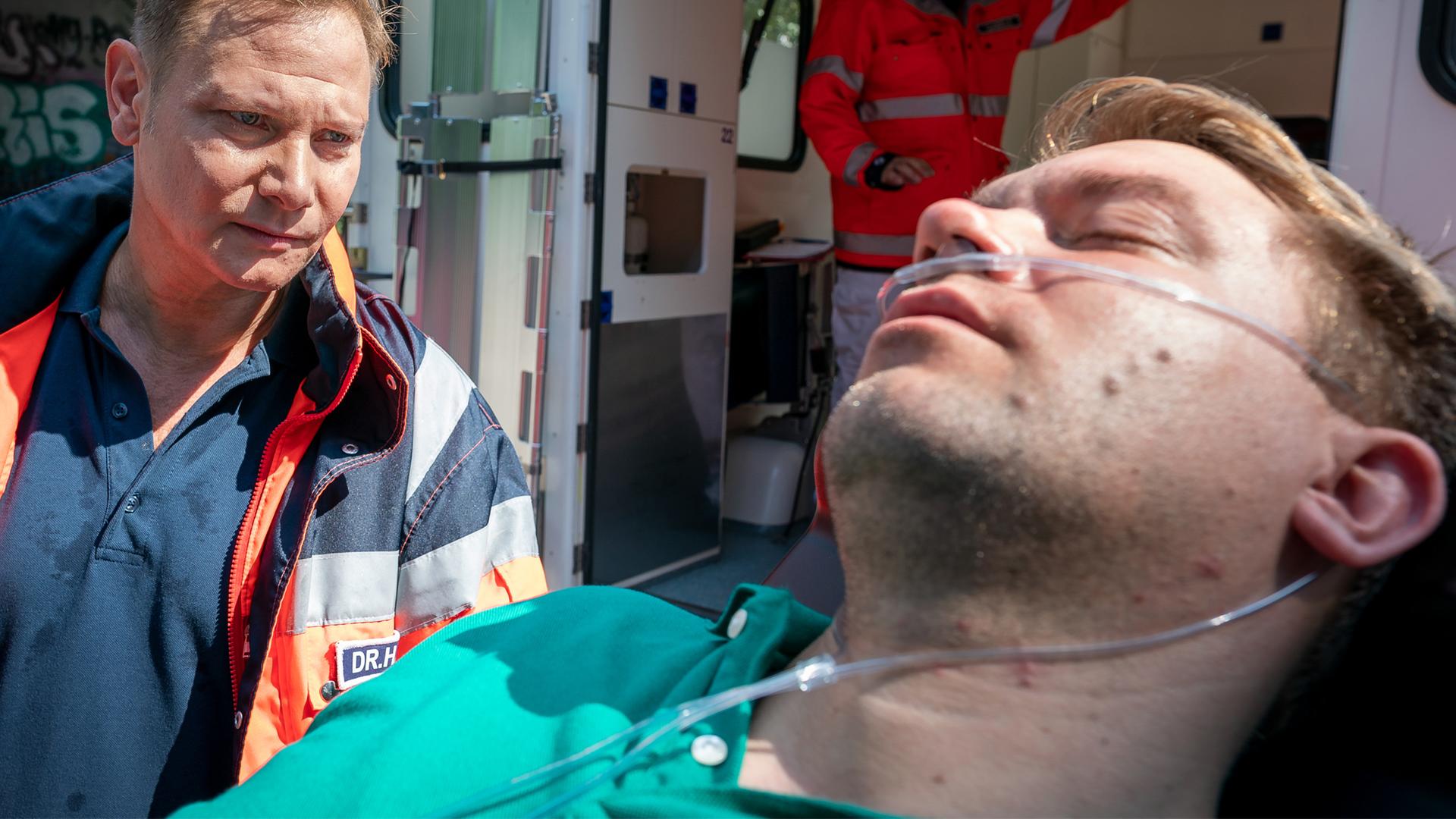 Dr. Haase (Fabian Harloff) steht neben dem Kopfende einer Patiententrage, auf der Peter Jüllich (David Korbmann) offenbar bewusstlos liegt. Haase blickt Jüllich nachdenklich an. Sie befinden sich hinter einem geöffneten Rettungswagen.