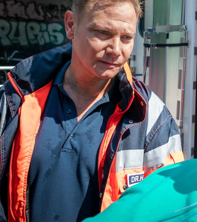 Dr. Haase (Fabian Harloff) steht neben dem Kopfende einer Patiententrage, auf der Peter Jüllich (David Korbmann) offenbar bewusstlos liegt. Haase blickt Jüllich nachdenklich an. Sie befinden sich hinter einem geöffneten Rettungswagen.